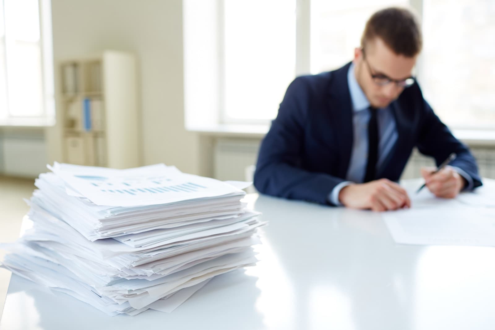 A tall stack of printed invoices on a desk with a manager reviewing paperwork behind